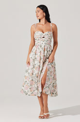 A woman is wearing a sleeveless dress with thin off-shoulder straps tied into bows on her shoulders. The dress has a fitted bodice with a twisted knot detail at the bust and a cutout below it. The skirt is long, flowy, and features a floral print with soft pastel colors. There is a slit on the front of the skirt revealing part of her leg. She is wearing open-toe sandals and has her dark hair down.